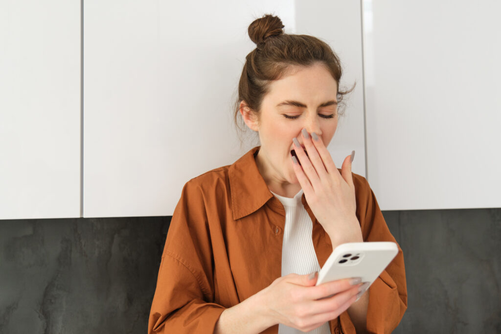 A bored young woman yawning while scrolling through videos on her phone, illustrating low audience retention on social media.